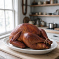 Whole hickory-smoked turkey with deep mahogany skin and legs tied with twine, on a white platter in a farmhouse kitchen